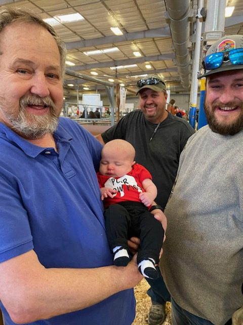 Steven Coston holding his granddaughter River in a PCPR Show Pigs shirt, with her father Adam and Dale Porter behind them at a livestock show